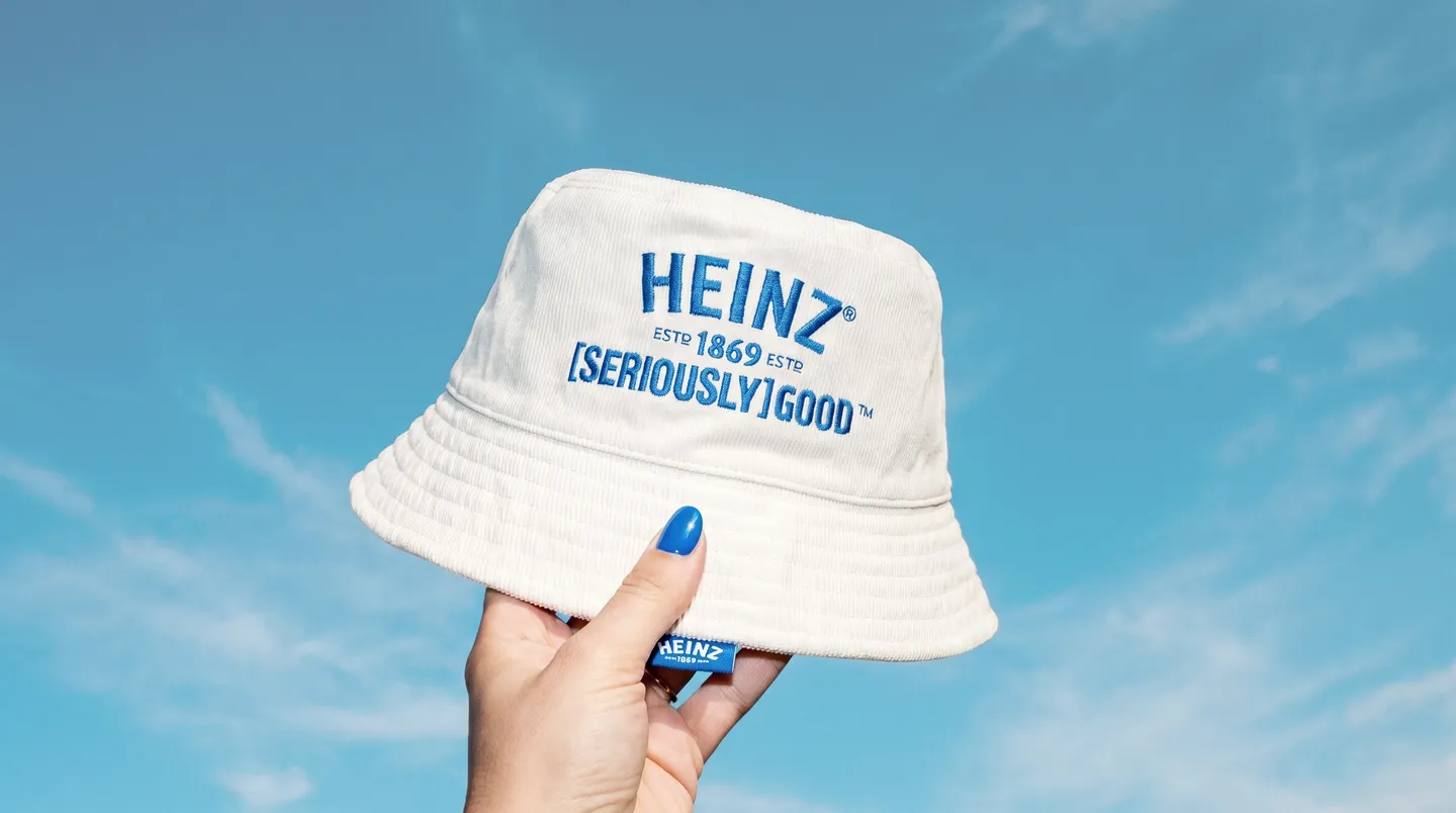 A hand holding a cream corduroy Heinz-branded bucket hat against a blue sky