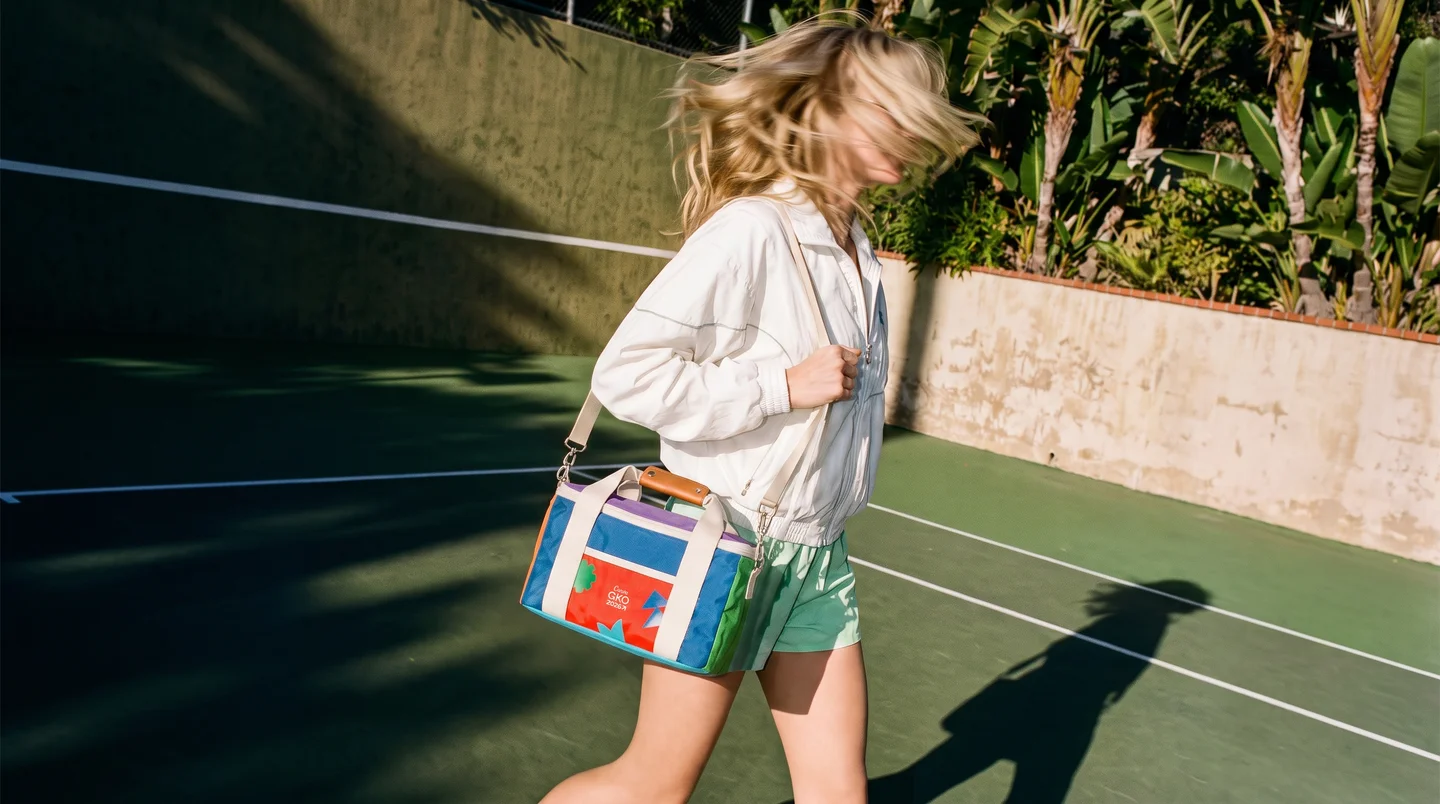 A woman walking across a tennis court with a colorful branded cooler bag over her shoulder