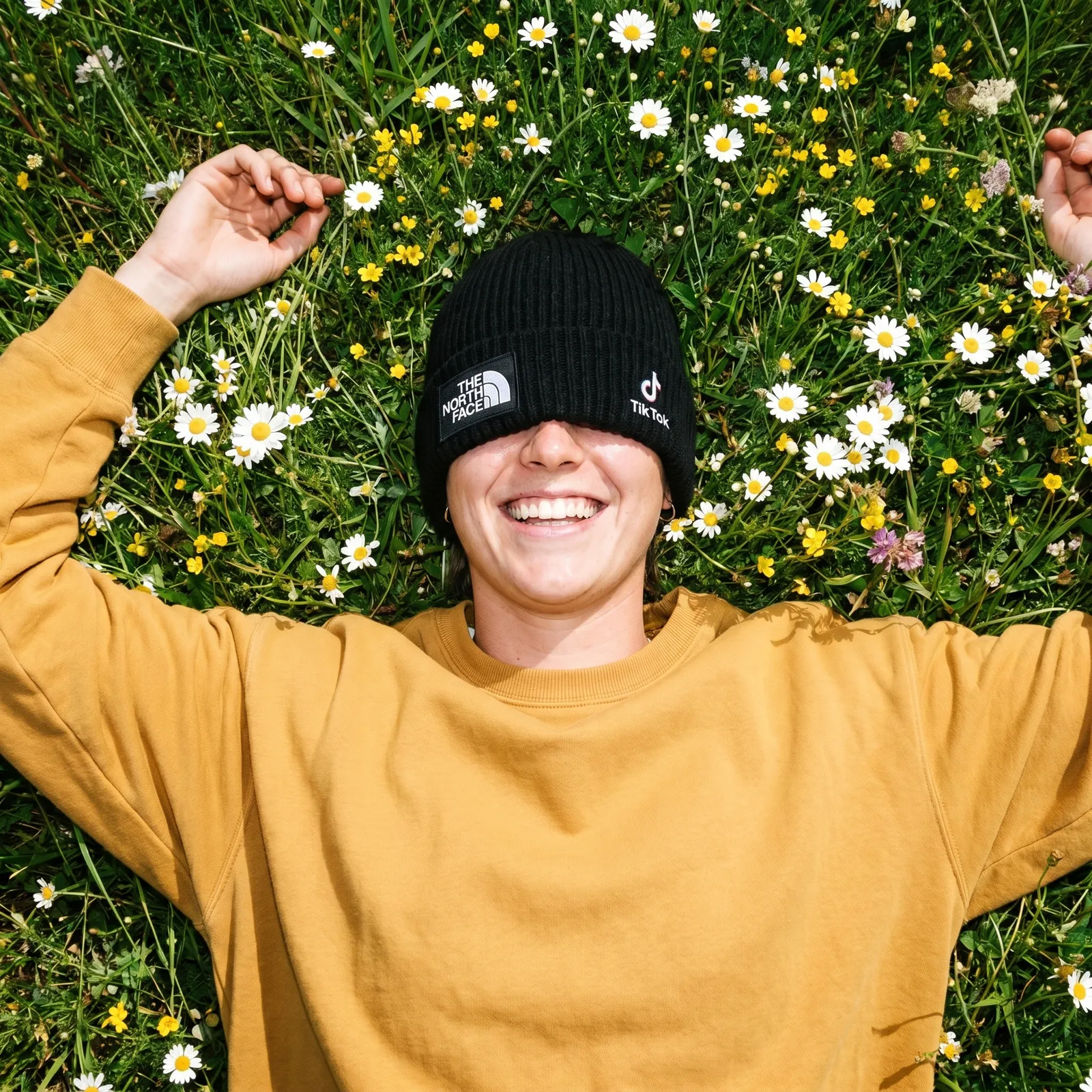 Smiling person in a flower meadow wearing a branded beanie